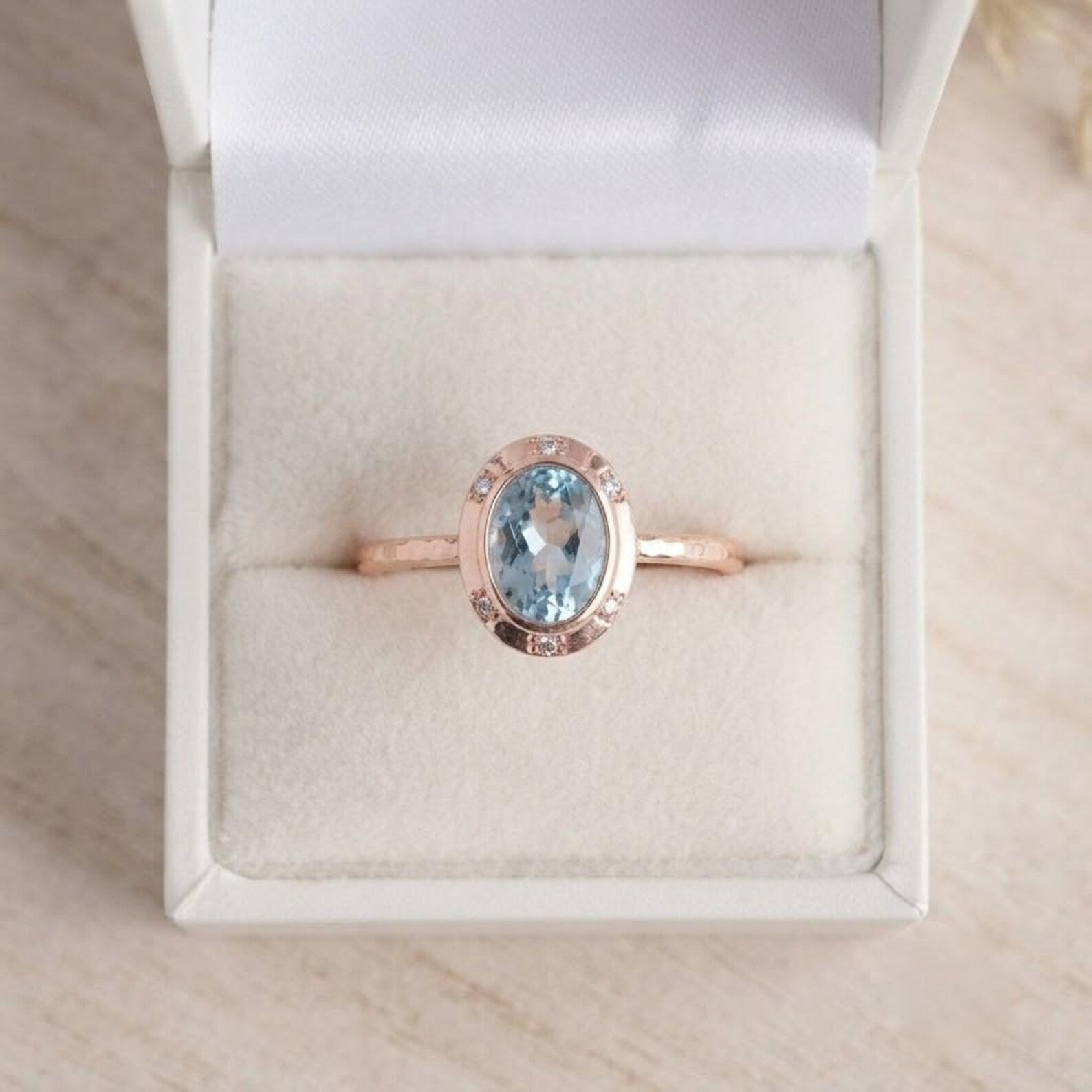 May include: A rose gold ring featuring a central oval-cut light blue gemstone surrounded by a halo of small clear stones. The band has a textured, hammered finish and is presented inside a white velvet-lined jewelry box with a hinged lid.