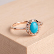 May include: A rose gold ring featuring a central oval-shaped turquoise cabochon stone. The setting is accented with three small, clear round-cut diamonds on each side of the turquoise, creating a symmetrical design on a thin, polished metal band.