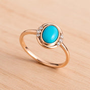May include: A rose gold ring featuring a central oval-shaped turquoise gemstone. The setting includes a smooth metal border and small, clear, round-cut accent stones embedded along the sides of the oval frame, creating a delicate look.