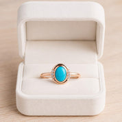 May include: A rose gold ring featuring a central oval-shaped turquoise gemstone. The setting includes three small, clear, round-cut accent stones on each side of the turquoise. The ring is displayed inside a cream-colored velvet jewelry box.