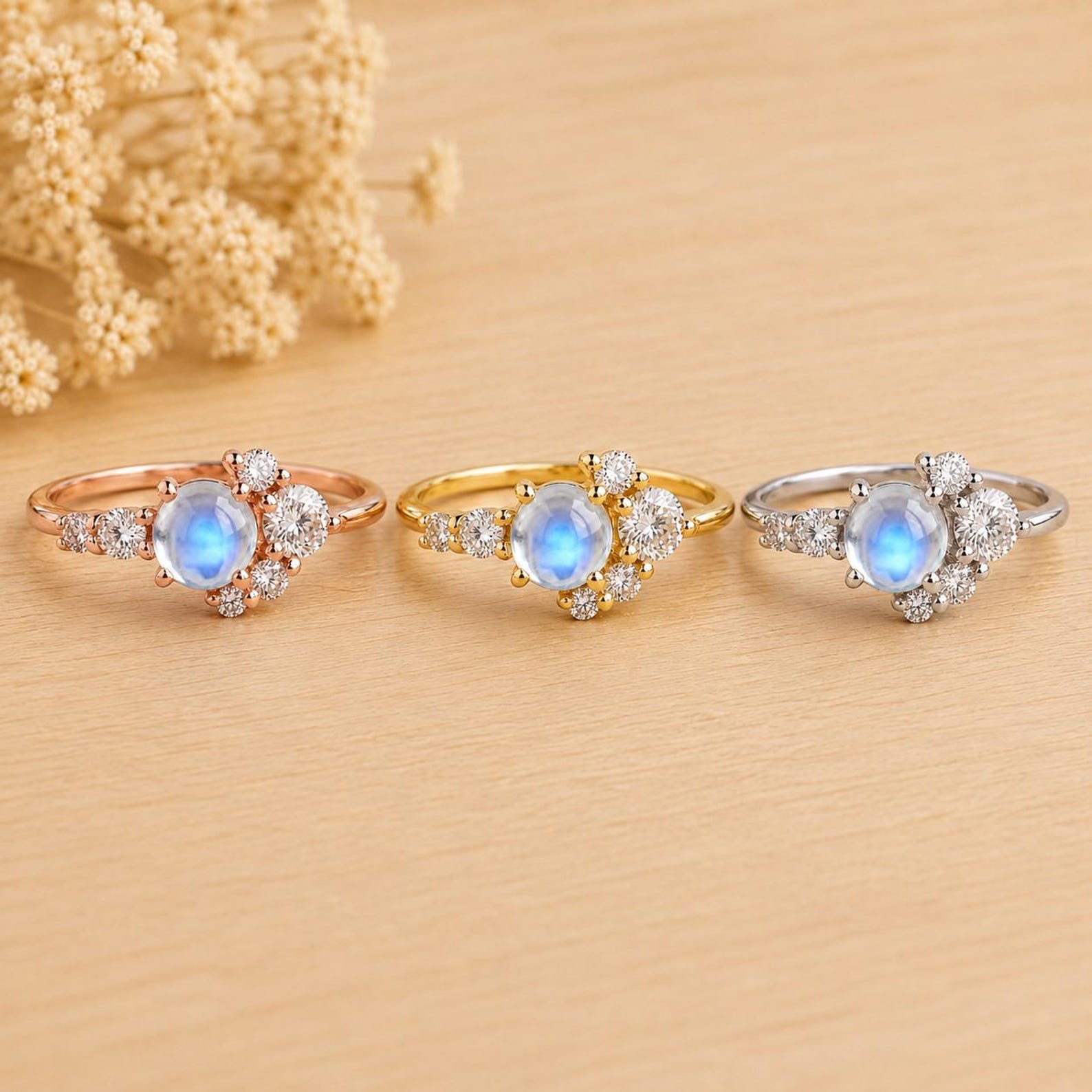 May include: Three rings featuring a central round cabochon moonstone with a blue glow, flanked by clusters of small round clear stones. The rings are presented in rose gold, yellow gold, and silver-toned metal finishes, arranged in a horizontal row.