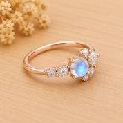 May include: A rose gold ring featuring a central round cabochon moonstone with a blue iridescent glow, flanked by a cluster of small, round, clear faceted stones set along the band in a prong setting, creating a delicate and balanced design.
