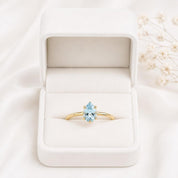 A gold ring with a blue gemstone is displayed in a white jewelry box on a white background.
