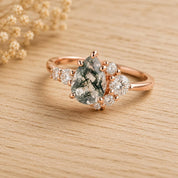 May include: A rose gold ring featuring a pear-shaped moss agate center stone with dark green dendritic inclusions. The band is accented with a cluster of round brilliant-cut white diamonds on both sides of the central gemstone setting.