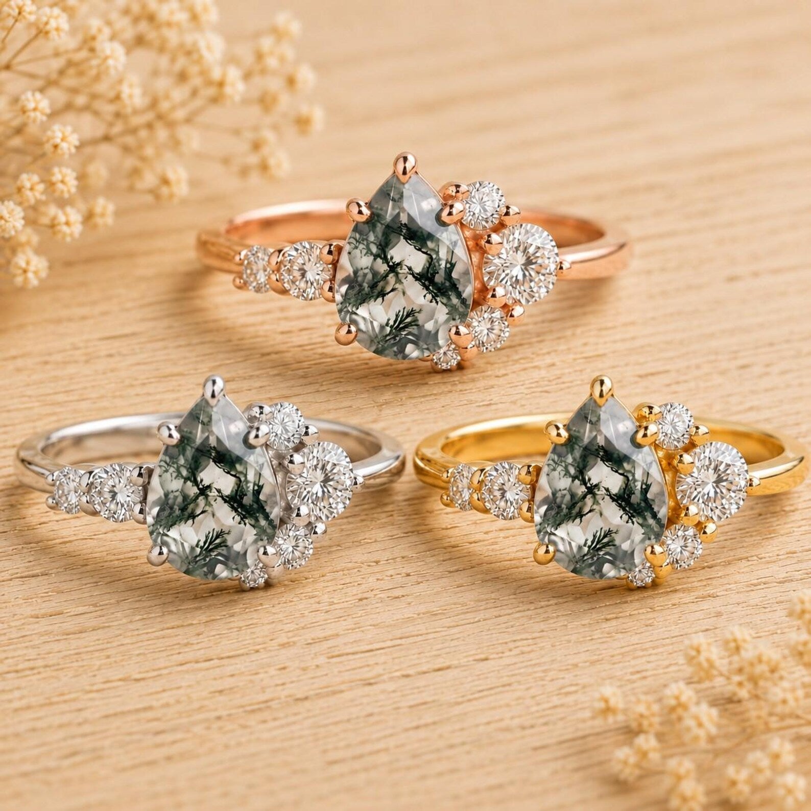 May include: Three rings featuring pear-shaped moss agate center stones with dendritic inclusions. Each ring is accented by a cluster of small round white stones on one side. The bands are crafted in rose gold, yellow gold, and silver-toned metal.