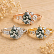 May include: Three rings featuring pear-shaped moss agate center stones with dendritic inclusions. Each ring is accented by a cluster of small round white stones on one side. The bands are crafted in rose gold, yellow gold, and silver-toned metal.