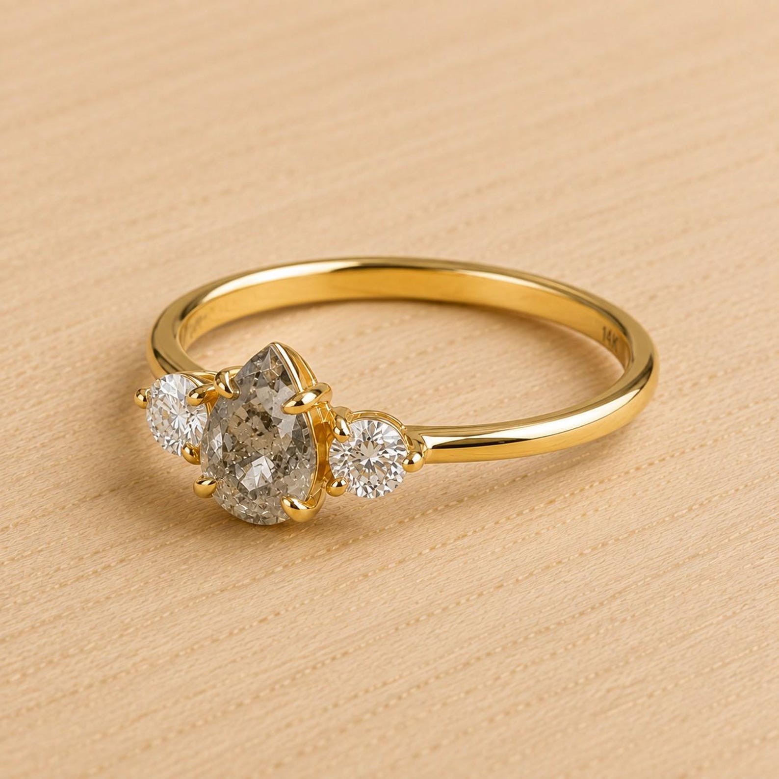 May include: A gold ring featuring a central pear-shaped salt and pepper diamond flanked by two smaller round brilliant-cut white diamonds. The band is polished yellow gold with a smooth, thin profile, showcasing a three-stone setting design.