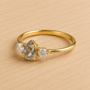 May include: A gold ring featuring a central pear-shaped salt and pepper diamond flanked by two smaller round brilliant-cut white diamonds. The band is polished yellow gold with a smooth, thin profile, showcasing a three-stone setting design.