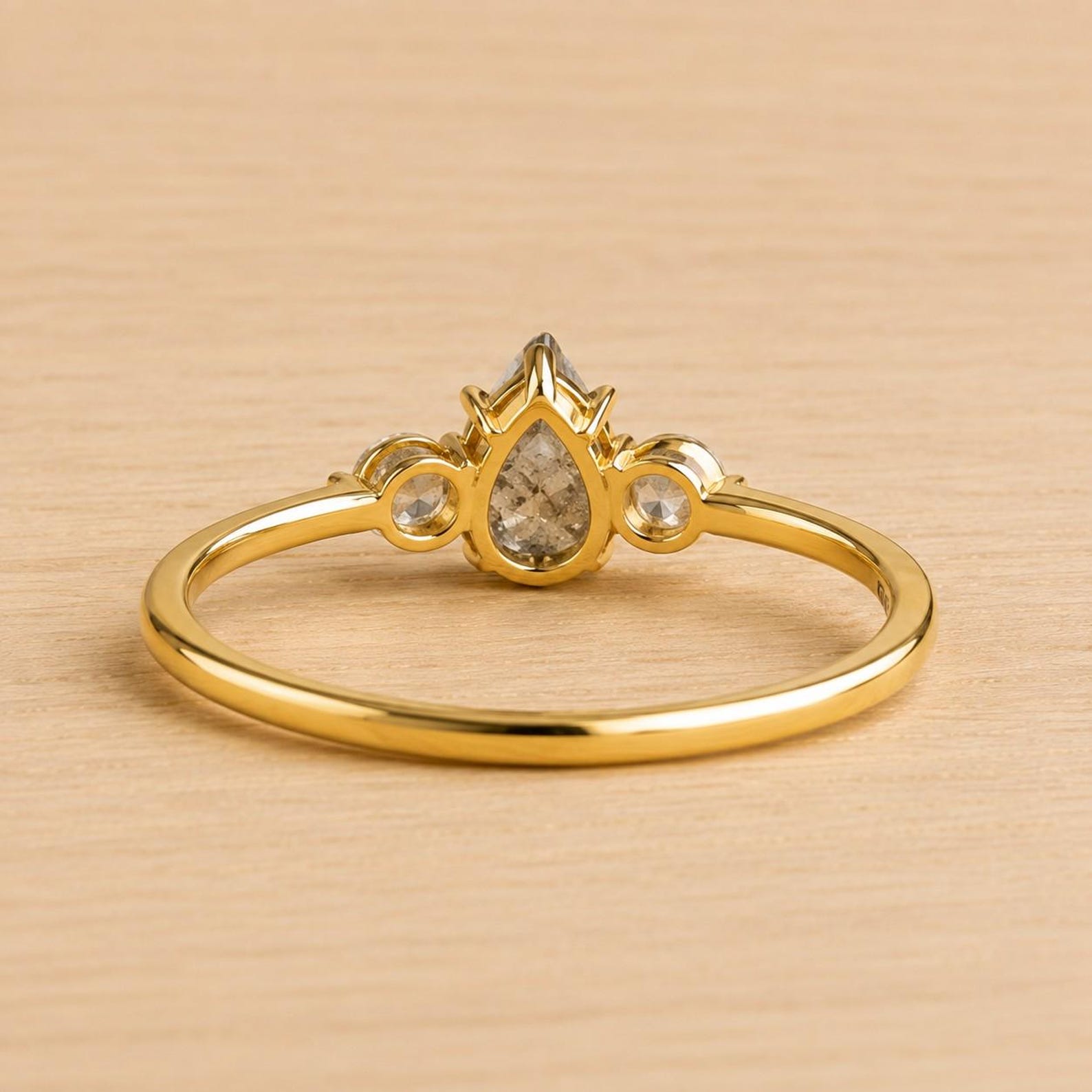 May include: A gold ring featuring a central pear-shaped stone set in a prong setting, flanked by two smaller round stones on either side. The band is thin and polished, showcasing a bright yellow gold finish that contrasts with the clear stones.
