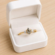 May include: A gold ring featuring a central pear-shaped salt and pepper diamond flanked by two small round brilliant-cut diamonds. The ring is presented inside a white hinged jewelry box with a soft white interior lining and a smooth exterior finish.