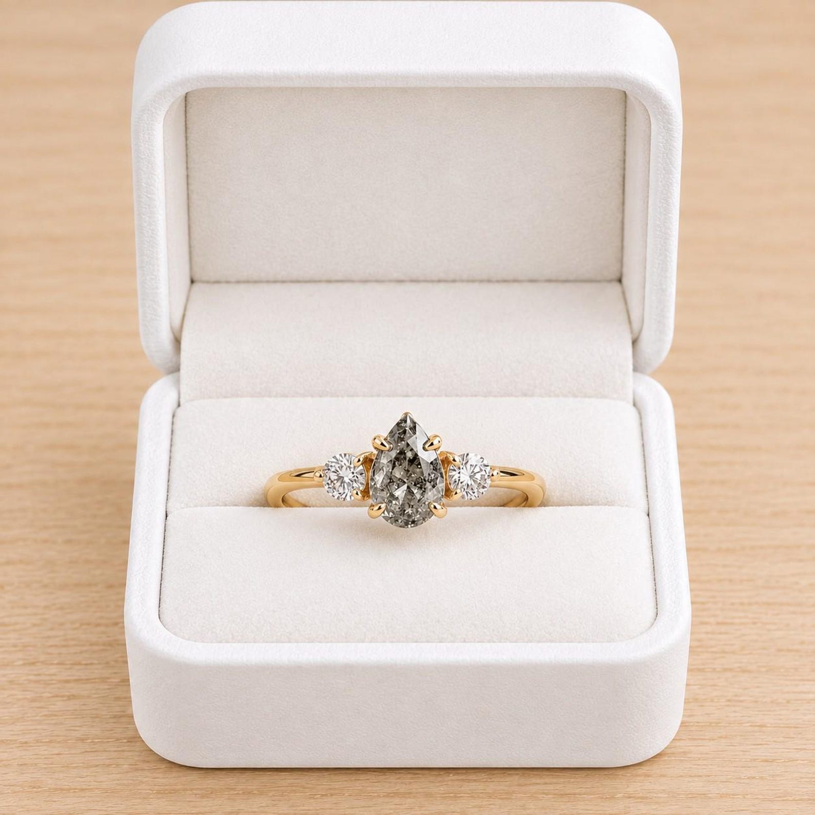 May include: A gold ring featuring a central pear-shaped salt and pepper diamond flanked by two smaller round brilliant-cut diamonds, all set in a prong setting, presented inside a white velvet-lined jewelry box with a hinged lid.