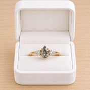 May include: A gold ring featuring a central pear-shaped salt and pepper diamond flanked by two smaller round brilliant-cut diamonds, all set in a prong setting, presented inside a white velvet-lined jewelry box with a hinged lid.