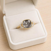 A gold ring with a large gray gemstone, placed in a white jewelry box.