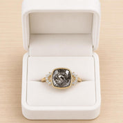 A white jewelry box containing a gold ring with a large gray gemstone, possibly a diamond, and smaller diamonds set in the band.
