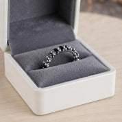A silver ring with black stones is displayed in a white jewelry box on a wooden surface.