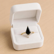 A white jewelry box containing a gold ring with a black gemstone, placed on a wooden surface.