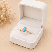 A white jewelry box containing a ring with a turquoise stone set in a gold band, placed on a wooden surface.